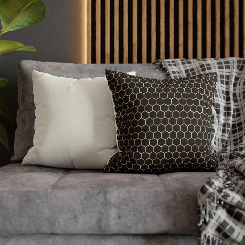 May include: Two square throw pillows are displayed. One pillow is solid off-white, while the other features a black background with a repeating gold-colored honeycomb pattern. Both pillows have a smooth, fabric texture and are square in shape.