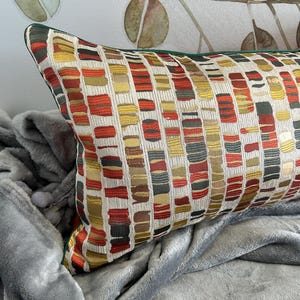 Puede incluir: Una almohada rectangular con un patr&oacute;n geom&eacute;trico en tonos rojos, amarillos, verdes y marrones. La almohada tiene un borde verde y est&aacute; sobre una manta gris.
