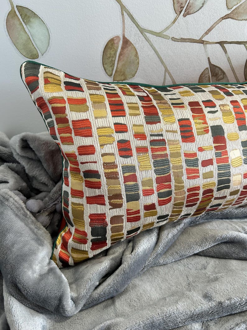Puede incluir: Una almohada rectangular con un patr&oacute;n geom&eacute;trico en tonos rojos, amarillos, verdes y marrones. La almohada tiene un borde verde y est&aacute; sobre una manta gris.