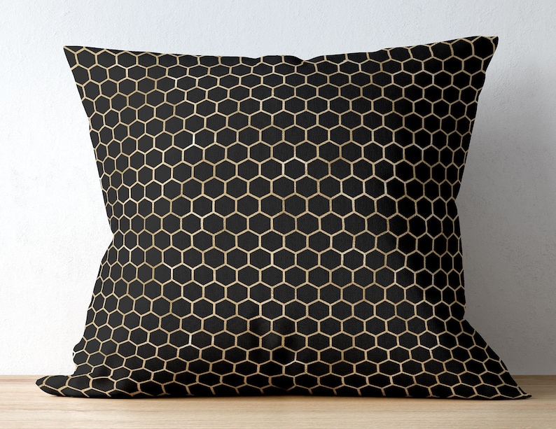 May include: A square throw pillow featuring a repeating geometric honeycomb pattern. The design consists of thin, metallic gold lines forming a grid of hexagons set against a solid, deep black background, creating a high-contrast visual effect.