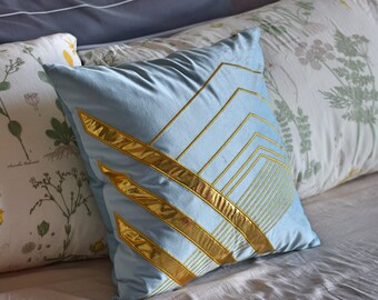 Puede incluir: Una almohada azul claro con bordado geométrico dorado. La almohada está sobre una cama con una sábana blanca y otras almohadas.