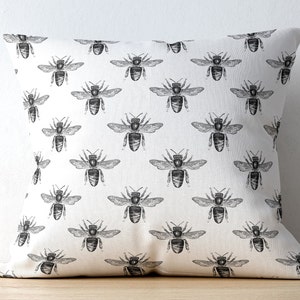 May include: A square throw pillow featuring a repeating pattern of black ink-style bee illustrations printed on a solid off-white fabric background. The design displays multiple bees oriented in various directions across the entire surface.