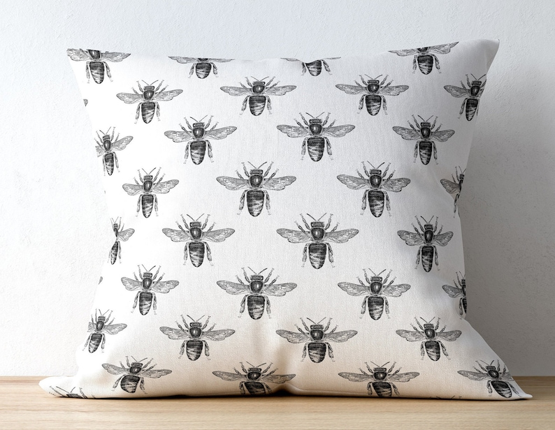 May include: A square throw pillow featuring a repeating pattern of black ink-style bee illustrations printed on a solid off-white fabric background. The design displays multiple bees oriented in various directions across the entire surface.