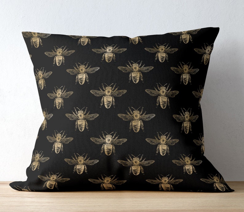 May include: A square throw pillow featuring a repeating pattern of gold-colored bees printed on a solid black fabric background. The bees are depicted in a detailed, sketch-like style with visible wings, antennae, and segmented bodies.
