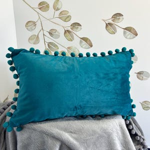 May include: Teal blue velvet cushion with a pom-pom trim. The cushion is sitting on a grey blanket.