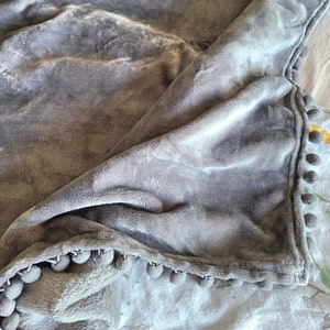 May include: A soft, gray throw blanket with a pom-pom trim. The blanket is folded and draped over a surface.