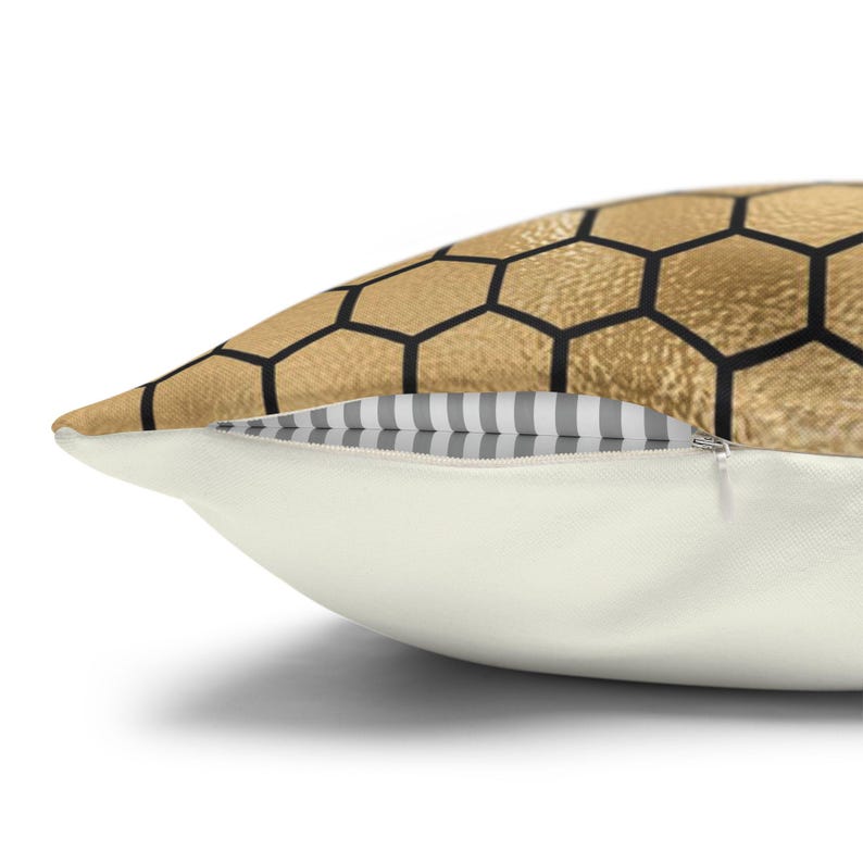 May include: A throw pillow featuring a gold-colored honeycomb pattern with black outlines on the top half and a solid off-white bottom half. A silver zipper is visible along the seam, revealing a grey and white striped fabric lining inside.