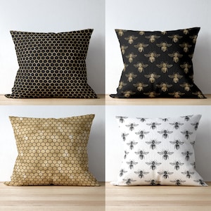 May include: A set of four square throw pillows. Two pillows feature a repeating honeycomb pattern in black and gold. One pillow has a black background with gold bee illustrations, and one pillow has a white background with black bee illustrations.