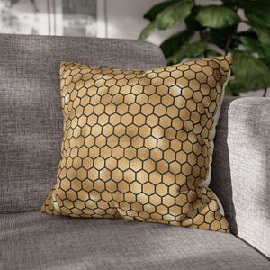 May include: A square throw pillow featuring a metallic gold surface with a repeating black hexagonal honeycomb pattern. The fabric has a textured finish, and the pillow is filled to maintain a plump, structured shape with defined edges.