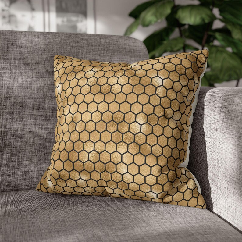 May include: A square throw pillow featuring a metallic gold surface with a repeating black hexagonal honeycomb pattern. The fabric has a textured finish, and the pillow is filled to maintain a plump, structured shape with defined edges.