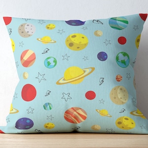May include: Square throw pillow with a light blue background and a repeating pattern of colorful planets, stars, and lightning bolts. The planets include Saturn, Earth, and other celestial bodies in shades of yellow, red, and blue.