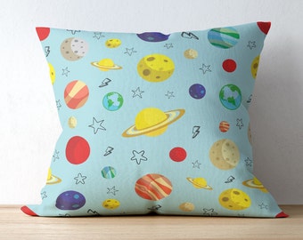 May include: Square throw pillow with a light blue background and a repeating pattern of colorful planets, stars, and lightning bolts. The planets include Saturn, Earth, and other celestial bodies in shades of yellow, red, and blue.