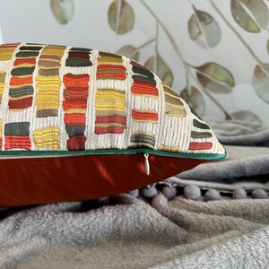 Puede incluir: Una almohada de tiro roja y dorada con un patr&oacute;n geom&eacute;trico y una cremallera oculta. La almohada est&aacute; sobre una manta gris y peluda.