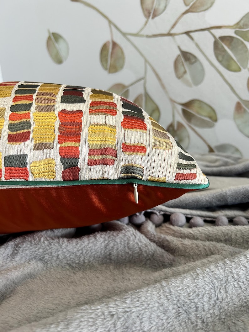 Puede incluir: Una almohada de tiro roja y dorada con un patr&oacute;n geom&eacute;trico y una cremallera oculta. La almohada est&aacute; sobre una manta gris y peluda.