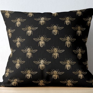 May include: A square throw pillow featuring a repeating pattern of gold-colored bees printed on a solid black fabric background. The bees are depicted in a detailed, sketch-like style with visible wings, antennae, and segmented bodies.