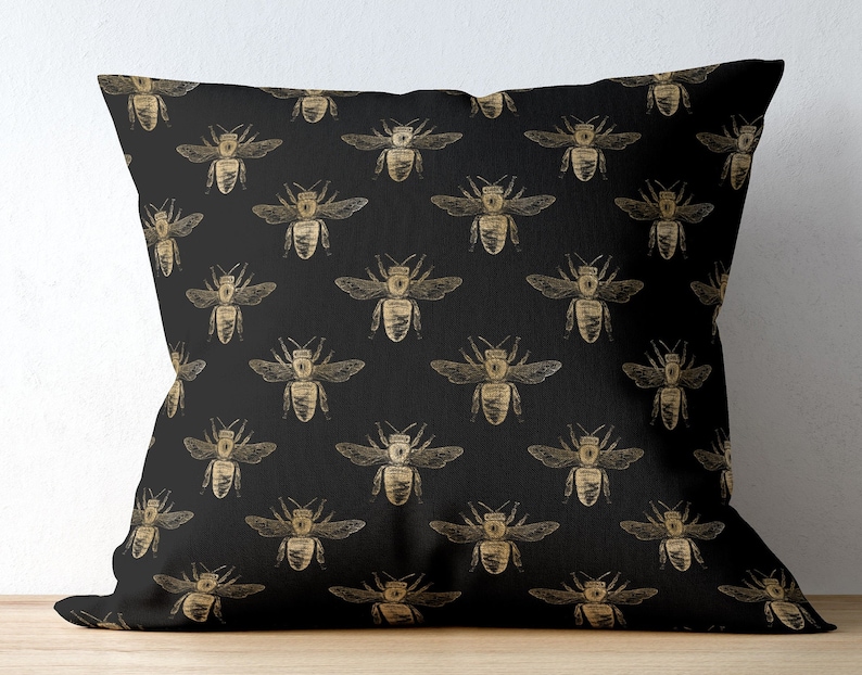 May include: A square throw pillow featuring a repeating pattern of gold-colored bees printed on a solid black fabric background. The bees are depicted in a detailed, sketch-like style with visible wings, antennae, and segmented bodies.