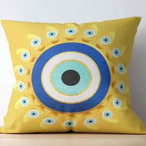 May include: A yellow throw pillow with a blue and white evil eye design. The evil eye is surrounded by a circle of smaller blue and white eyes.