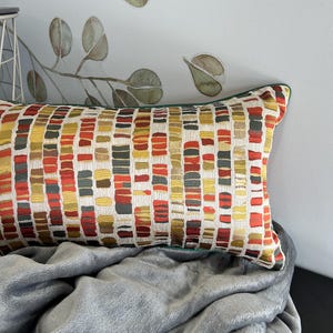 Puede incluir: Una almohada lumbar rectangular con un patr&oacute;n geom&eacute;trico en tonos de rojo, naranja, amarillo y verde. La almohada tiene un borde verde y est&aacute; sobre una manta gris.