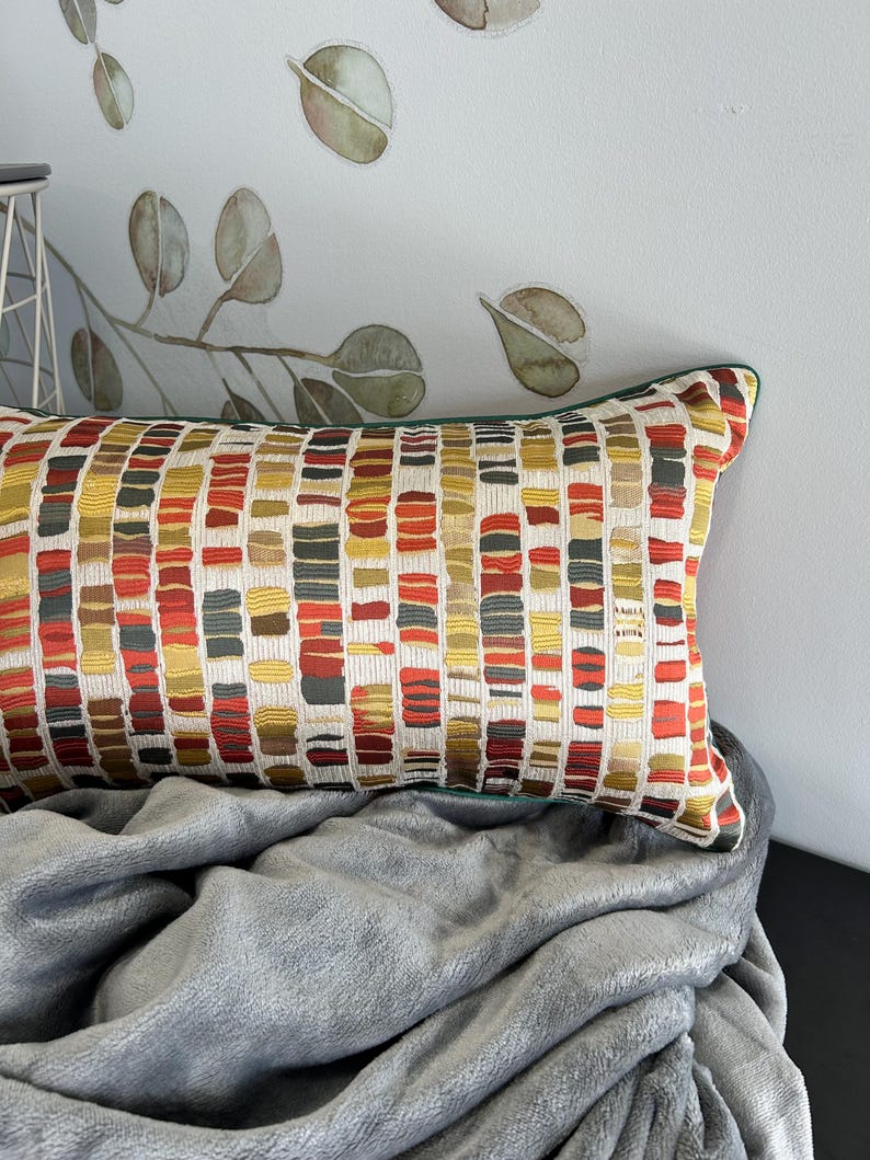 Puede incluir: Una almohada lumbar rectangular con un patr&oacute;n geom&eacute;trico en tonos de rojo, naranja, amarillo y verde. La almohada tiene un borde verde y est&aacute; sobre una manta gris.