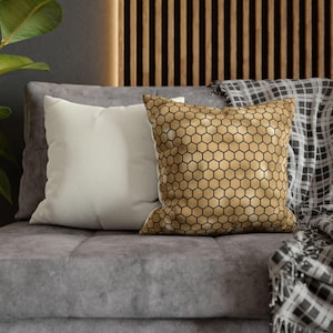 May include: Two square throw pillows placed on a gray sofa. One pillow is solid off-white, while the other features a metallic gold surface with a repeating black honeycomb or hexagonal geometric pattern printed across the entire front face.