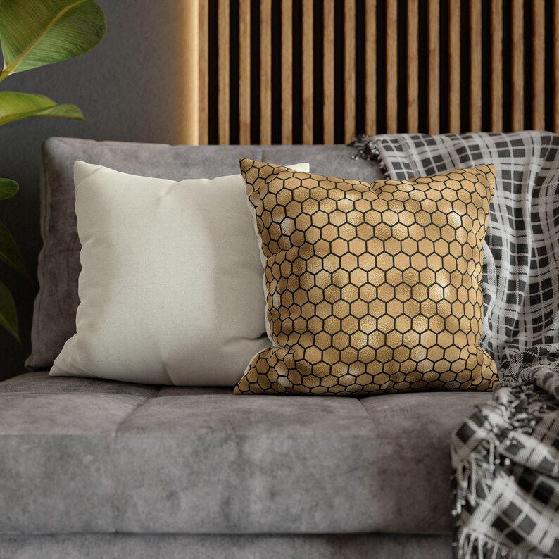 May include: Two square throw pillows placed on a gray sofa. One pillow is solid off-white, while the other features a metallic gold surface with a repeating black honeycomb or hexagonal geometric pattern printed across the entire front face.