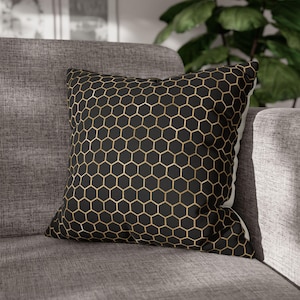 May include: A square throw pillow featuring a black fabric base with a repeating gold-colored honeycomb or hexagonal geometric pattern printed across the front surface. The pillow has a structured shape and is finished with clean, straight edges.