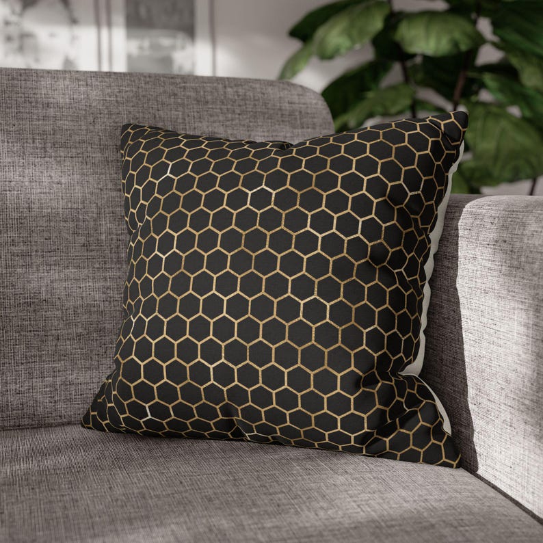 May include: A square throw pillow featuring a black fabric base with a repeating gold-colored honeycomb or hexagonal geometric pattern printed across the front surface. The pillow has a structured shape and is finished with clean, straight edges.