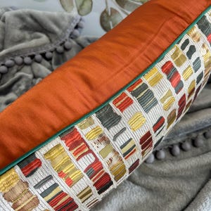 Puede incluir: Una almohada decorativa con un patr&oacute;n geom&eacute;trico en tonos de rojo, amarillo, verde y blanco. La almohada tiene un respaldo de color &oacute;xido y un ribete verde.