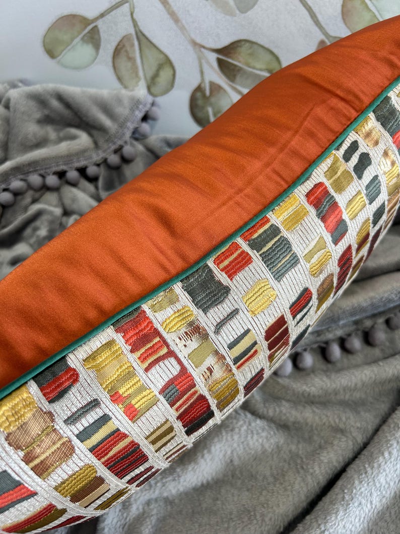 Puede incluir: Una almohada decorativa con un patr&oacute;n geom&eacute;trico en tonos de rojo, amarillo, verde y blanco. La almohada tiene un respaldo de color &oacute;xido y un ribete verde.