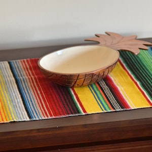 May include: A colourful striped table runner with a pineapple-shaped bowl on top. The runner has yellow, red, green, blue and white stripes.