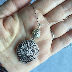May include: A silver locket necklace with a filigree design and a small pearl-like bead. The locket is open and the inside is visible.