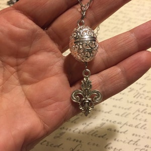 May include: A silver necklace with a filigree pendant and a fleur-de-lis charm. The pendant is a hollow sphere with intricate designs.