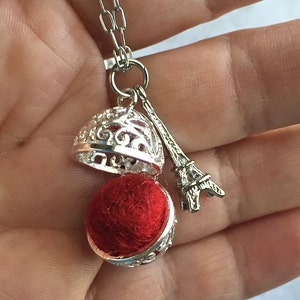 Op de afbeelding: Een zilveren ketting met een rode vilten bal bedel in een kooi-achtige hanger. De hanger is bevestigd aan een zilveren Eiffeltoren bedel.