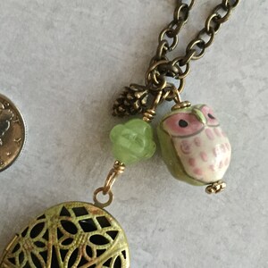 Puede incluir: Un collar con un medallón verde y dorado con un delicado diseño de filigrana. El medallón tiene un pequeño cristal en el centro. El collar también incluye una cuenta de vidrio verde, un pequeño amuleto de piña de pino dorado y un amuleto de búho rosa y blanco.