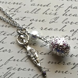 May include: A silver necklace featuring three charms: a stylized figure, a filigree orb with a red interior, and a small, dark purple bead. The necklace is set against a backdrop of handwritten script.