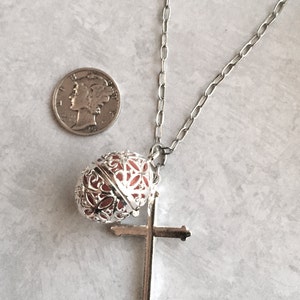 May include: A silver chain necklace with a silver cross pendant and a silver orb with a red interior. The orb is decorated with intricate floral designs.