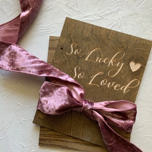 May include: A wooden sign with the text "So Lucky So Loved" in cursive script, with a small heart cutout. The sign is tied with a dusty rose velvet ribbon.