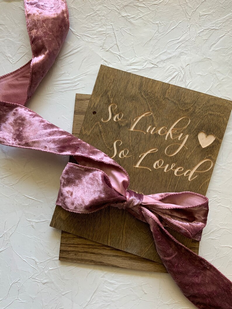 May include: A wooden sign with the text "So Lucky So Loved" in cursive script, with a small heart cutout. The sign is tied with a dusty rose velvet ribbon.
