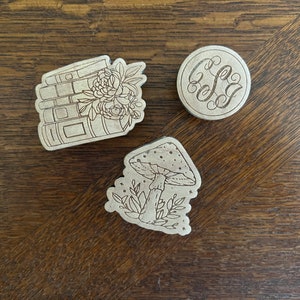 May include: Three wooden laser-cut stickers. One sticker features a stack of books with flowers, another has a mushroom with leaves, and the last one has a monogram with the letters "S&J".