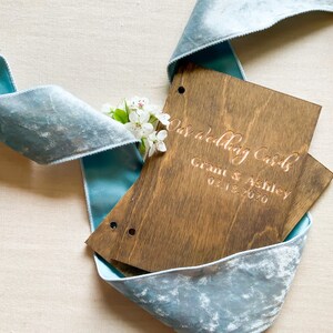 May include: A wooden box with the text "Our Wedding Cards" and "Grant & Ashley 02.18.2020" engraved on the front. The box is wrapped in a light blue velvet ribbon.