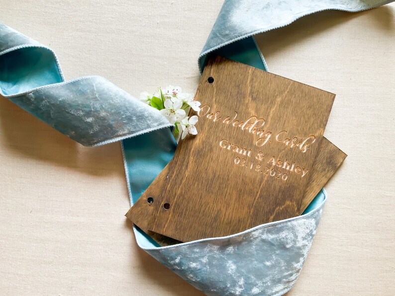 May include: A wooden box with the text "Our Wedding Cards" and "Grant & Ashley 02.18.2020" engraved on the front. The box is wrapped in a light blue velvet ribbon.