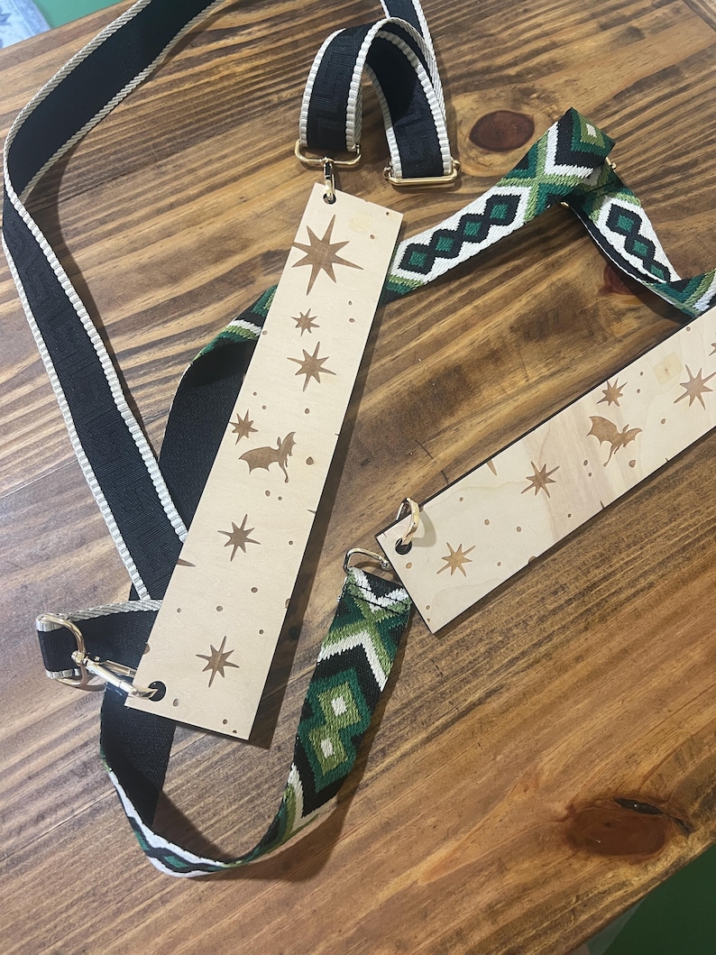 May include: A collection of bag straps and wooden tags on a wooden surface. One strap is black with a white stripe, and another is green and white with a geometric pattern. The wooden tags have star and bat designs.