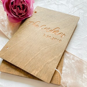 May include: A wooden guest book with the text "The Coolers 3.30.2019" engraved in gold lettering. The book is closed and sitting on a white surface with a pink velvet ribbon.