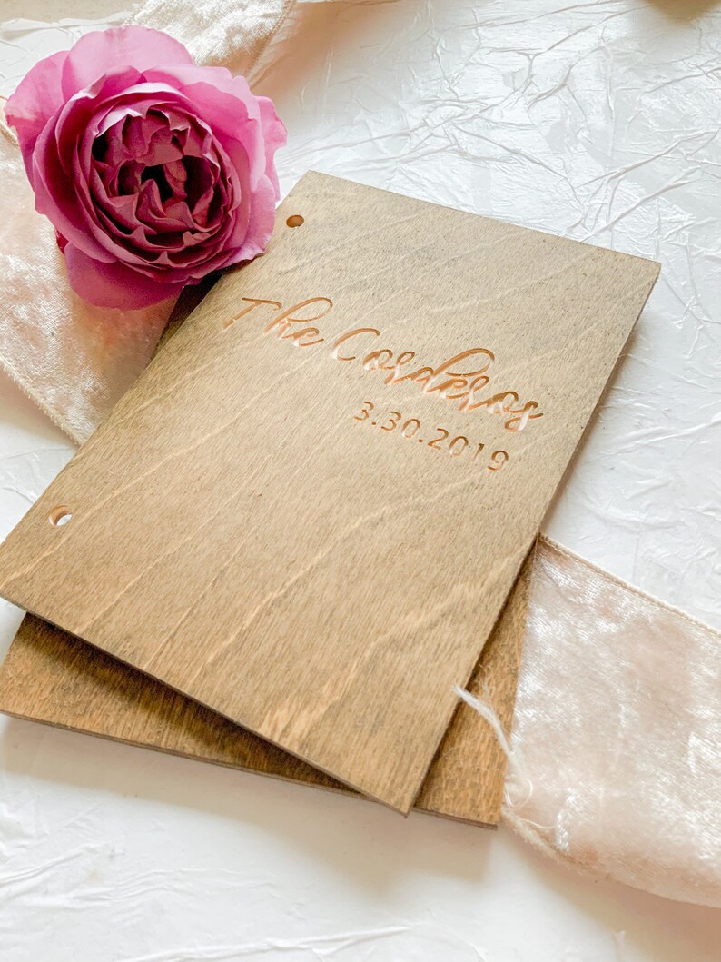 May include: A wooden guest book with the text "The Coolers 3.30.2019" engraved in gold lettering. The book is closed and sitting on a white surface with a pink velvet ribbon.