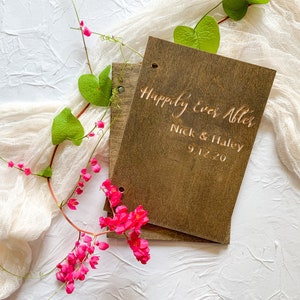 May include: A wooden guest book with the text "Happily Ever After" engraved on the front. The names "Nick & Haley" and the date "9.1.20" are also engraved on the front. The book is surrounded by pink flowers and green leaves.