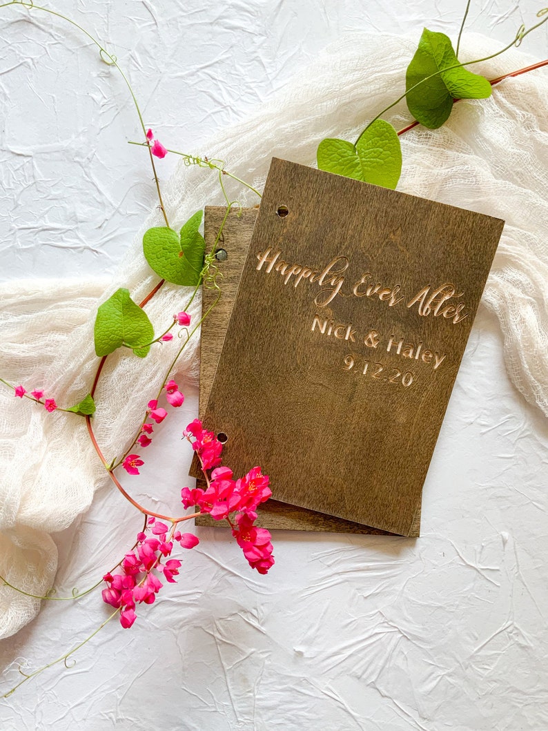 May include: A wooden guest book with the text "Happily Ever After" engraved on the front. The names "Nick & Haley" and the date "9.1.20" are also engraved on the front. The book is surrounded by pink flowers and green leaves.
