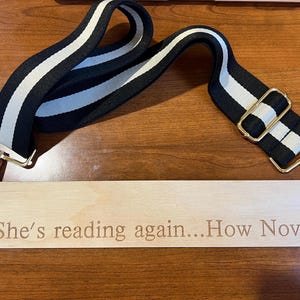 May include: A black and white striped bag strap with gold-colored metal clasps and a wooden tag. The tag is engraved with the phrase "She's reading again...How Novel". The strap is made of a durable fabric.