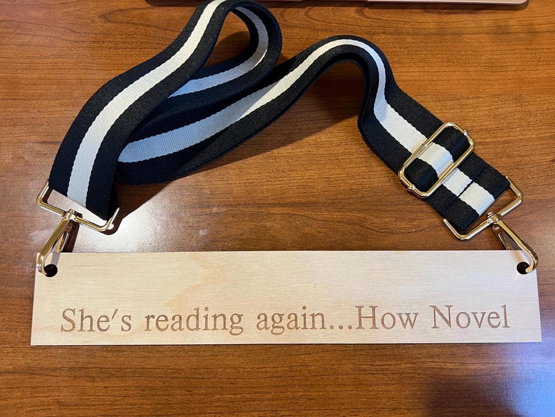 May include: A black and white striped bag strap with gold-colored metal clasps and a wooden tag. The tag is engraved with the phrase "She's reading again...How Novel". The strap is made of a durable fabric.