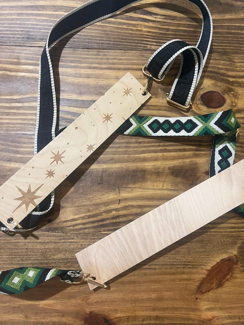 May include: Wooden bag strap with starburst and star designs, paired with a black and white striped strap and a green and white patterned strap. The straps have gold-colored metal hardware and are displayed on a wooden surface.