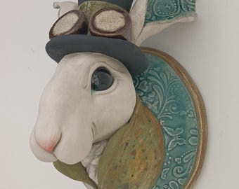 May include: A whimsical ceramic rabbit head sculpture. The rabbit wears a black top hat and goggles. The sculpture is mounted on a teal-colored, ornate, oval-shaped plaque with a gold-colored border. The rabbit's fur is white, with a pink nose.